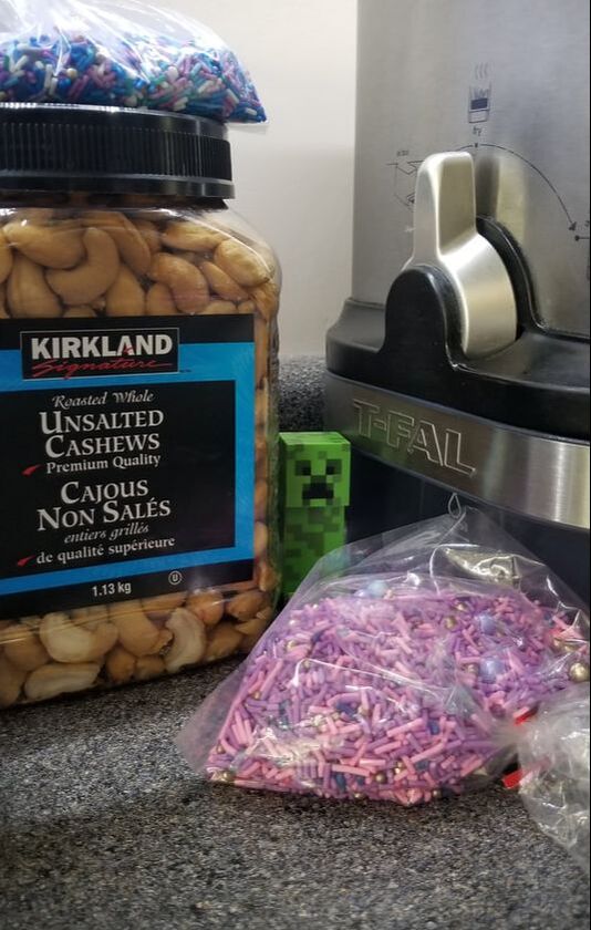 Photo of a small minecraft creeper toy between a deep fryer, a bulk bag of cake sprinkles, and a container of pecans on a grey kitchen counter